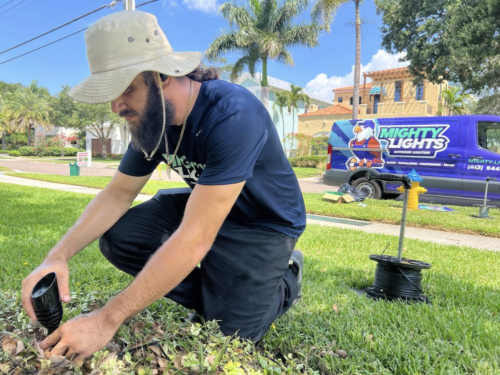 Mighty Lights technician installing a landscape lighting fixture on a Tampa Bay lawn with the wrapped service van parked in the background